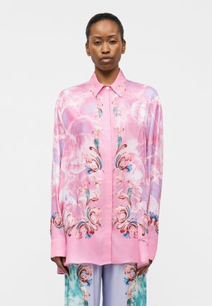 Frau trägt ein locker sitzendes rosa Seidenhemd mit kunstvollen blauen und orangefarbenen Blumenmustern, passende Hosen, steht vor weißem Hintergrund.