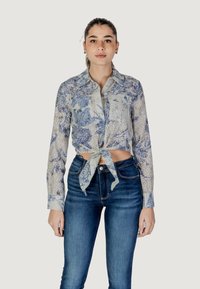 Bloemige overhemdo met lange mouwen in lichtblauw en gebroken wit. Voorzien van een strik vooraan en twee borstzakken. Dit wordt gecombineerd met donkerblauwe jeans.