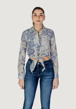 Bloemige overhemdo met lange mouwen in lichtblauw en gebroken wit. Voorzien van een strik vooraan en twee borstzakken. Dit wordt gecombineerd met donkerblauwe jeans.