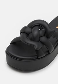 Sandales plates noires avec une bride en cuir nouée, présentant une texture lisse, un bout arrondi et une semelle épaisse pour plus de hauteur.
