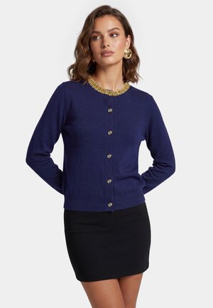 Donna che indossa un cardigan blu navy con bottoni dorati e scollo decorato, abbinato a una gonna nera, in piedi con le mani dietro la schiena.