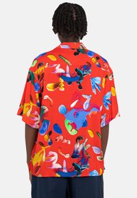 Short-sleeve red shirt featuring a colorful abstract pattern with various shapes, made of smooth fabric. Back view shows a relaxed fit.