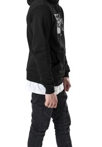 Felpa nera con cappuccio e stampa grafica frontale, abbinata a jeans scuri con pannelli testurizzati e una camicia bianca stratificata sotto.