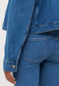 Denim jakna u srednje opranom plavu, s opuštenim krojem, dugmadima na manžetama i detaljima na stražnjem šavu, u kombinaciji s uskim plavim trapericama.