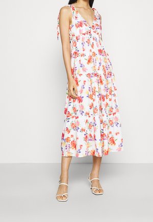Frau trägt ein weißes ärmelloses Midi-Kleid mit Blumenmuster und Bindebändern sowie weiße Riemchen-Sandaletten mit Absatz und steht vor einem schlichten Hintergrund.