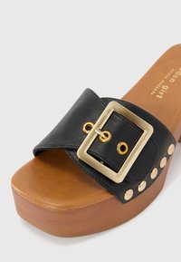 Sandal slide em couro preto com uma tira larga, fivela em hardware dourado e tachas douradas decorativas ao longo do salto. Sola de plataforma texturizada em tom castanho.