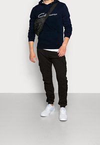 Hombre con sudadera navy con logotipo en cursiva, pantalones cargo negros, zapatillas blancas y bolso cruzado negro, de pie en un suelo gris contra una pared blanca.