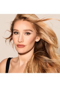 Donna con lunghi capelli biondi mossi, che indossa una canotta nera. Le sue caratteristiche includono sopracciglia definite, rossetto neutro e pelle radiosa.