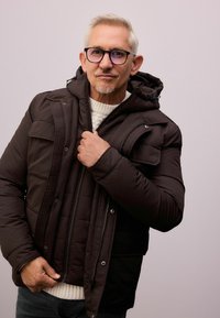 Gepolsterte braune Jacke mit Kapuze, ausgestattet mit einem Reißverschluss und Druckknöpfen, getragen über einem cremefarbenen Strickpullover. Sichtbares Detail des strukturierten Stoffes.