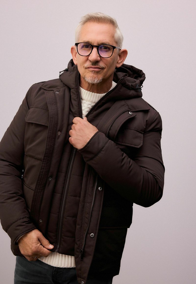 Gepolsterte braune Jacke mit Kapuze, ausgestattet mit einem Reißverschluss und Druckknöpfen, getragen über einem cremefarbenen Strickpullover. Sichtbares Detail des strukturierten Stoffes.