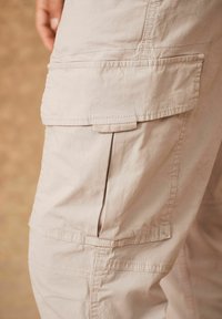Pantaloni cargo beige chiaro con numerose tasche laterali, vestibilità comoda e dettagli di cucitura visibili. Presentano un tessuto morbido e strutturato.