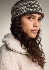 Un bandeau en tricot marron et crème avec un motif texturé. Porté avec une veste en polaire beige dotée d'une fermeture éclair et d'un col haut.