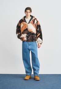 Giacca oversize e soffice con motivo astratto marrone, arancione e crema, abbinata a jeans a gamba larga azzurri chiaro e zoccoli color cuoio.