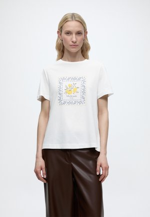 Vrouw draagt een wit shirt met korte mouwen met een gele citroengrafiek en een blauwe bloemenrand, gecombineerd met donkerbruine leren broek.