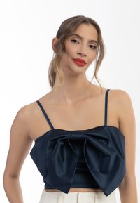 Námořnická saténová crop top s tenkými ramínky a velkým mašlí vpředu. Měkká textura, strukturovaný tvar a přiléhavý design akcentují siluetu.