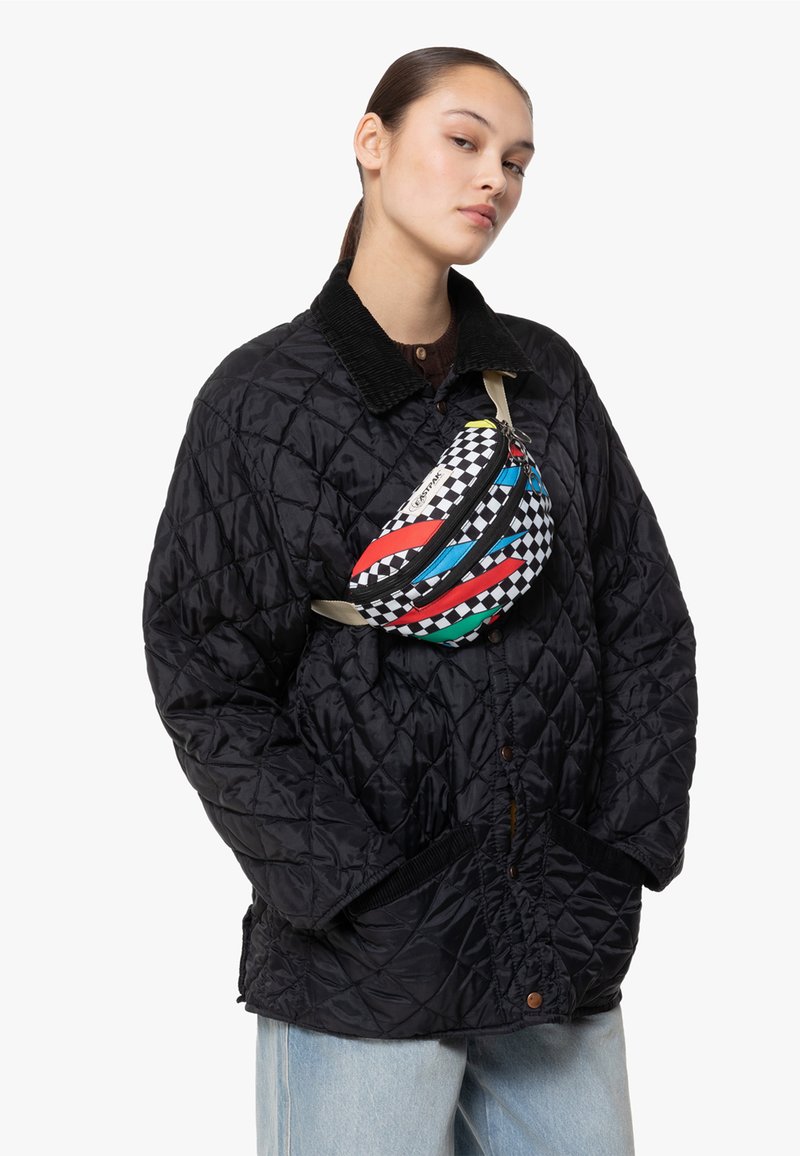 Quiltad svart jacka med en kordflosshals. Färgglad, mönstrad midjeväska med rutig design och dragkedjor bärs över bröstet.