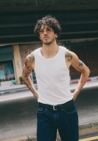 Canottiera di cotone bianca abbinata a jeans denim blu scuro e una cintura nera. Sono in vista tatuaggi sui bracci e occhiali con montatura dorata.