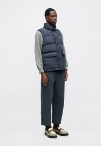 Chaleco acolchado azul marino con cuello alto, dos bolsillos frontales, llevado sobre una camisa de manga larga gris clara, combinado con pantalones oscuros y zapatos distintivos.