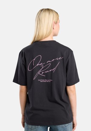 Camiseta de algodón negra con texto cursivo rosa en la parte trasera, que dice "Una ronda más" junto con un lema gráfico abajo. Mangas cortas.