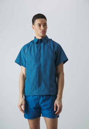 Chemise courte à manches courtes bleu canard avec une finition texturée, col montant et fermeture éclair à l'avant. Associée à un short bleu canard assorti, le tout en matière légère.