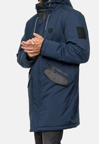 Parka matelassée bleu marine avec capuche, accents noirs, poche latérale zippée et détails de poignets élastiques. Présente un logo sur la poitrine.