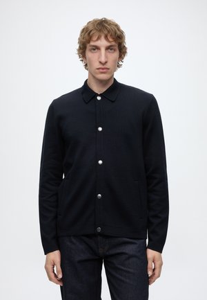 Homme aux cheveux bouclés portant une veste noire boutonnée et un jean foncé, debout devant un fond clair uni.
