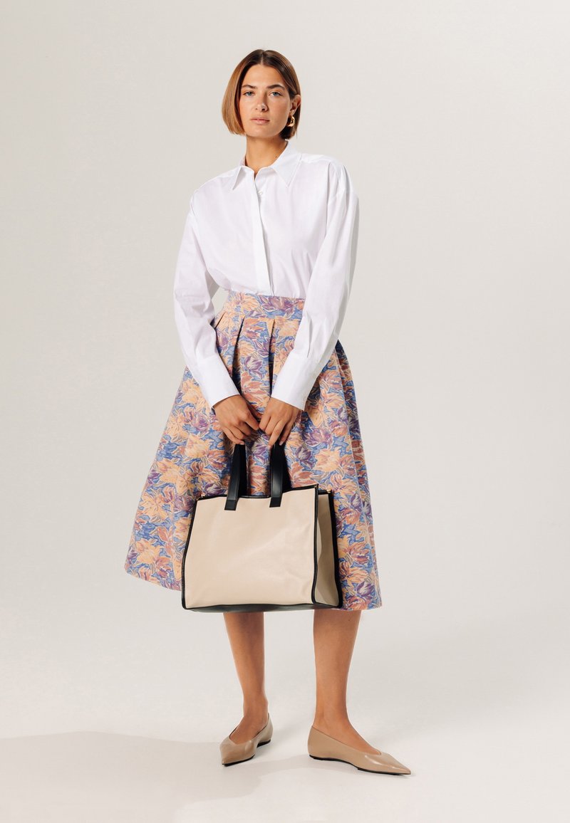 Sac fourre-tout en cuir beige avec des accents noirs, jupe à motifs floraux dans des tons bleus et roses, chemise blanche à boutons, et chaussures plates beiges.