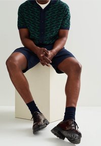 Muž sedící na bílém bloku, oblečený v zelené vzorované košili, námořnických šortkách, tmavých ponožkách a hnědých kožených šněrovacích botách.