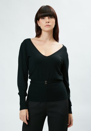 Zwarte trui met V-hals, lange mouwen en een aansluitende taille die wordt geaccentueerd door een smalle riem. Zachte textuur en minimalistisch design.