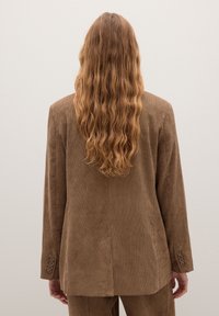 Bruine corduroy blazer met verticale strepen, met een enkele split aan de achterkant en knoopmanchetten, vanachter gezien met lang, golvend haar.