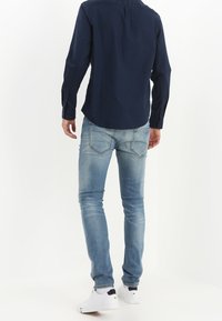 Maglia a maniche lunghe blu marina con polsini bottonati, abbinata a jeans denim azzurri e sneakers bianche. La vestibilità slim mette in risalto la forma e il design.