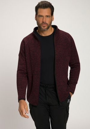 Chaqueta burdeos con cremallera, fabricada con un tejido texturizado, cuello alto y detalles en negro, combinada con una camisa y pantalones negros.
