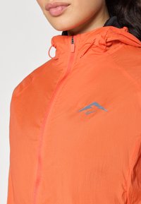 Lättviktig orange jacka med dragkedja fram, huva och en blå logotyp-accent på bröstet. Har en slät, texturerad tyg.