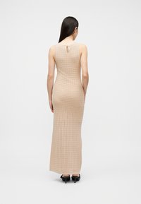 Γυναίκα με μακριά σκούρα μαλλιά, φοράει αμάνικο, μπεζ, υφασμάτινο μάξι φόρεμα και μαύρες γόβες, στέκεται με την πλάτη προς τα εμπρός σε λευκό φόντο.