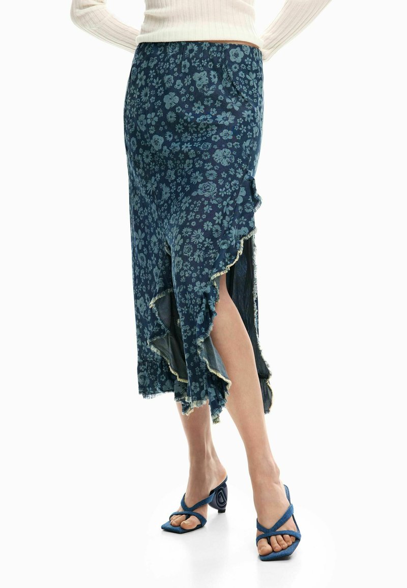 Kvinde iført en marineblå blomstret midinederdel med asymmetrisk flæsekant og blå hælede sandaler, stående med krydsede ben.