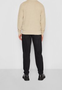 Mężczyzna ubrany w beżowy sweter, czarne dżinsy i czarne buty stoi tyłem do gładkiej, jasnoszarej ściany.