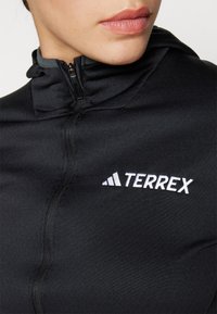 Čierna zipsová bunda vyrobená z hladkého, elastického materiálu; má šedý golier a biele logo "TERREX" na prednej strane.