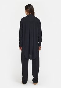 Chemise noire à manches longues avec un ourlet incurvé, fabriquée dans un tissu lisse. Le dos présente un détail de pli et un col classique.