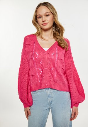 Cardigan roz tricotat cu decolteu în V, având închideri cu nasturi, modele texturate și mâneci bufante. Asociat cu blugi albaștri deschiși.