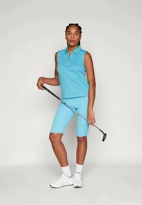 Γυναίκα σε αμάνικο μπλε γκολφ κοστούμι, κρατώντας μια γκολφ ρακέτα, στέκεται με αυτοπεποίθηση σε ένα απλό ανοιχτό φόντο.