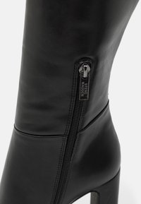 Bota alta de cuero negro hasta la rodilla con cremallera lateral, tacón alto grueso y textura suave; cuenta con un detalle de costura.