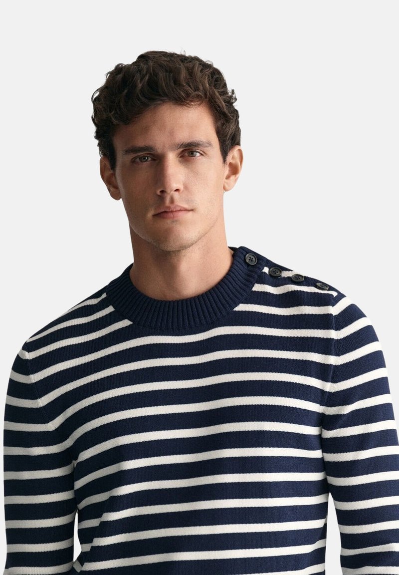 Hombre con cabello corto, rizado y oscuro, que lleva un suéter a rayas azul marino y blanco con botones en el hombro, expresión neutral, fondo claro y liso.