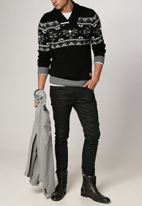 Pull en maille noir avec des motifs géométriques, accents gris, superposé sur une chemise blanche. Pantalons slim noirs et bottes noires, tenant une veste grise.