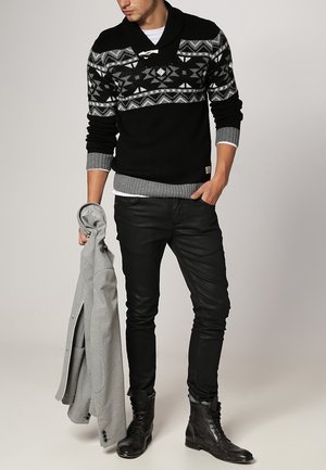 Schwarzer Strickpullover mit geometrischen Mustern, grauen Akzenten, über einem weißen Hemd. Schmale schwarze Hosen und schwarze Stiefel, eine graue Jacke haltend.