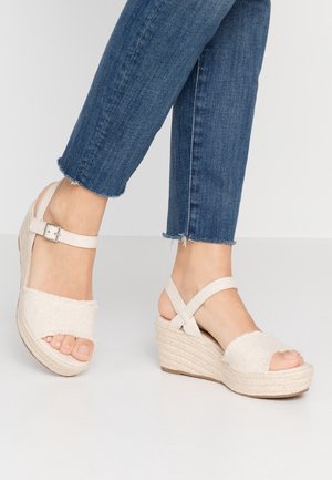 Pieds portant des sandales compensées en tissu beige avec brides à la cheville, associées à un jean bleu court à ourlet effiloché sur un fond uni.
