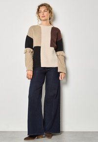 Maglione lavorato a maglia in beige, nero e marrone con pannelli testurizzati e polsini arricciati, abbinato a jeans scuri a gamba larga e stivaletti.