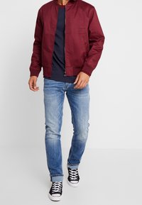 Burgundy bomber jacket with a zip front and ribbed cuffs, worn over a navy T-shirt and paired with light blue jeans and black sneakers.
