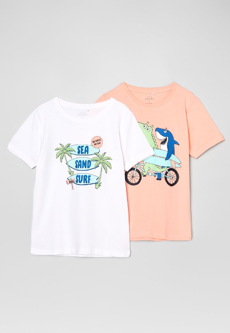 Dos camisetas: una blanca con el texto "SEA SAND SURF" y palmeras; otra melocotón con dibujos de dinosaurios en bicicleta. Mangas cortas, tejido de algodón.