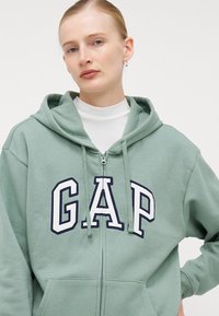 Világoszöld cipzáras kapucnis pulóver, nagy fehér és sötétkék "GAP" logóval, húzózsinórral ellátott kapucnival és elülső zsebekkel. Pamut keverék anyag.