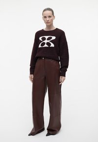 Bordeaux gekleurde gebreide trui met een wit geometrisch ontwerp, gecombineerd met hoog-waisted brede leren broek in bruin en puntige zwarte schoenen.